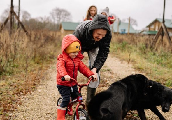 Dad helps son ride a bike on a path with family and dog, capturing a joyful outdoor moment.