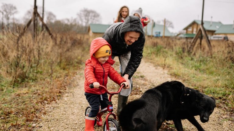 Dad helps son ride a bike on a path with family and dog, capturing a joyful outdoor moment.